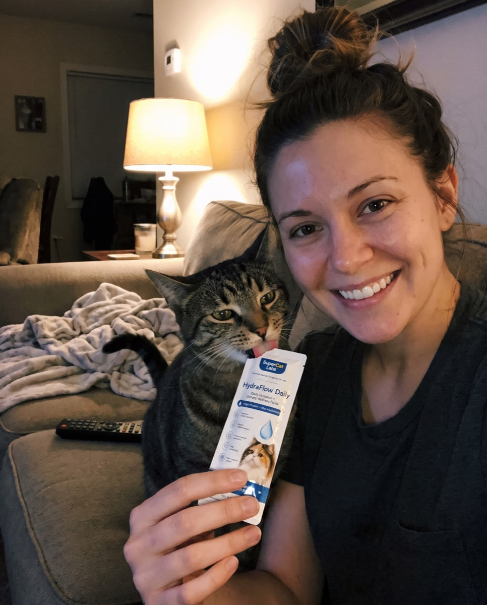 A smiling cat parent taking a couch selfie while her tabby licks a HydraFlow Daily sachet